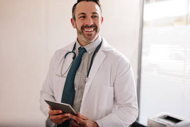 Smiling mature male doctor with a digital tablet in his office. Successful medical professional at his clinic. - JLPSF16620