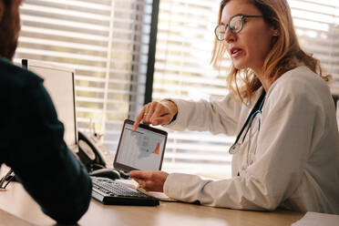 Ärztin spricht mit ihrem Patienten über Testergebnisse auf einem Tablet. Arzt und Patient sprechen über ein medizinisches Testergebnis mit einem männlichen Patienten, der auf ihrem Schreibtisch sitzt. - JLPSF16629