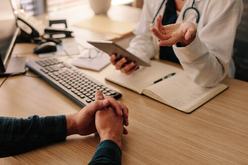 Eine Ärztin berät einen männlichen Patienten an ihrem Schreibtisch in der Klinik. Ausschnitt einer Ärztin, die ein digitales Tablet hält und mit ihrem Patienten in ihrem Büro spricht. - JLPSF16630