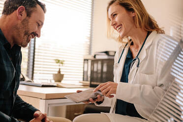 Eine Ärztin zeigt einem männlichen Patienten gute Testergebnisse. Eine lächelnde Ärztin teilt ihrem Patienten gute Testergebnisse mit. - JLPSF16632