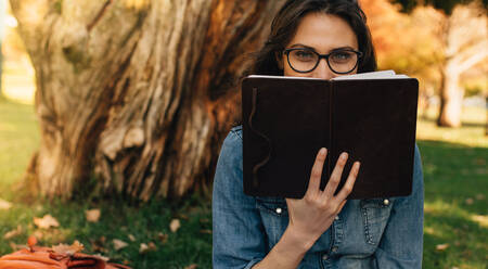 Frau schaut über ein Buch in der Hand. Studentin mit Brille schaut in die Kamera, während sie im Freien im Park ein Buch liest. - JLPSF16635