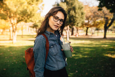 Porträt einer schönen Frau, die im Park steht und Fruchtsaft trinkt. Sie trägt ein Jeanshemd und eine Brille und hat eine Tasche auf der Schulter, aus der sie Saft trinkt. - JLPSF16637