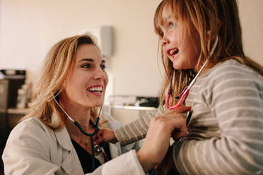 Lächelnde junge Kinderärztin, die mit einem Mädchen in ihrer Klinik spielt. Freundliche Ärztin und junges Mädchen, die sich gegenseitig mit einem Stethoskop untersuchen. - JLPSF16642