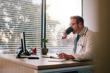 Reifer männlicher Arzt, der an seinem Schreibtisch Kaffee trinkt. Der Arzt arbeitet an seinem Schreibtisch und trinkt Kaffee. - JLPSF16657