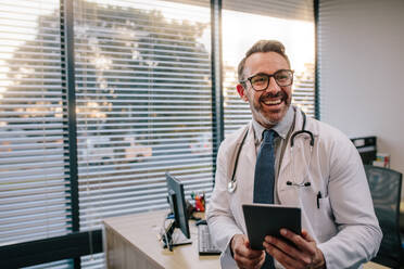 Smiling mature male doctor with digital tablet in his office. Friendly medical professional with tablet computer in clinic. - JLPSF16662