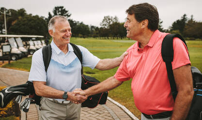 Two mature men are shaking hands and smiling when meeting on a golf course. Senior golfers at the golf course greeting each other with a handshake. - JLPSF16663