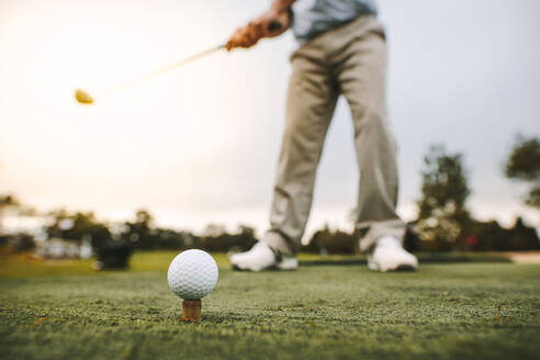 Golf ball on tee with man blurred at back about to make a shot at golf course. Focus on golf ball on tee with male golfer taking a shot. - JLPSF16671