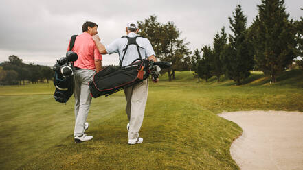 Rear view of two senior golf players walking together in the golf course with their golf bags. Senior golfers walking towards the next hole. - JLPSF16678