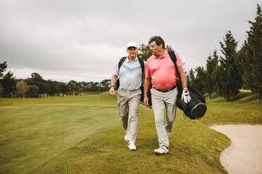 Two senior friends walking through the lawn with golf equipment and talking. Senior golf players walking together on the golf course. - JLPSF16679