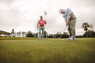 Low angle view of senior golfer on putting green about to take the shot. Male golf player putting on green with second male player in the background holding the flag. - JLPSF16680