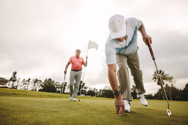 Ein älterer Golfspieler holt einen Golfball aus dem Loch, während ein zweiter Spieler im Hintergrund die Fahne hält. Ältere Golfspieler spielen eine Partie auf dem Golfplatz. - JLPSF16681