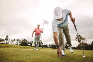 Senior golf player picking golf ball out of hole with second player in the background holding the flag. Senior golf players playing a game on the golf course. - JLPSF16681