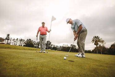 Männlicher Golfspieler bei einem Schlag auf dem Putting Green mit einem zweiten Spieler im Hintergrund, der die Fahne hält. Professionelle Golfspieler spielen zusammen auf dem Platz. - JLPSF16682