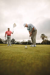 Professionelle Golfer spielen zusammen auf dem Golfplatz. Ein älterer Golfer macht einen Schlag auf dem Grün, während ein anderer Spieler im Hintergrund steht und die Flagge hält. - JLPSF16683