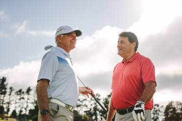 Two senior golf players with golf clubs standing together and talking between the game on a sunny day. Professional golfers having chat between the game on golf course. - JLPSF16689