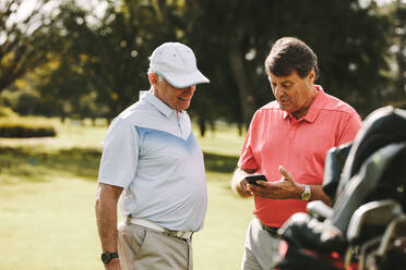 Senior man taking break from golfing and using mobile phone. Mature man showing the score to his friend after the game on golf course. - JLPSF16691