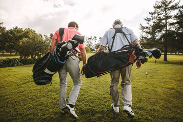 Rear view of two senior golf players walking together in the golf course with their golf bags. Senior golfers walking out of the course after the game. - JLPSF16694