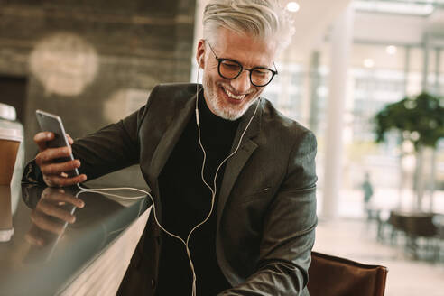 Lächelnder reifer Mann im Geschäftsanzug, der mit Kopfhörern im Café sitzt und ein Mobiltelefon in der Hand hält. Geschäftsmann, der mit Kopfhörern im Café telefoniert. - JLPSF16697