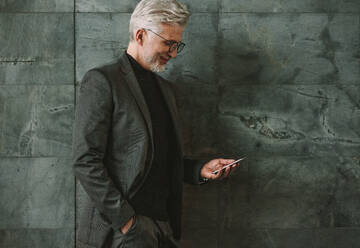 Älterer Geschäftsmann lehnt an einer Wand und schaut auf sein Handy. Älterer Geschäftsmann benutzt sein Handy auf grauem Hintergrund. - JLPSF16704