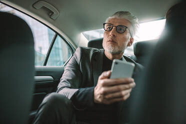 Älterer Geschäftsmann sitzt auf dem Rücksitz eines Taxis mit Mobiltelefon. - JLPSF16711