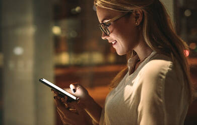 Seitenansicht einer glücklichen jungen Frau, die auf ihr Smartphone schaut und lächelt. Geschäftsfrau liest Textnachricht im Büro. - JLPSF16719