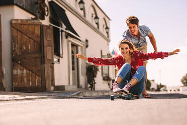 Smiling woman sitting on a skateboard sliding down an empty street. Man helping his girlfriend ride on a skateboard pushing her from behind. - JLPSF16722