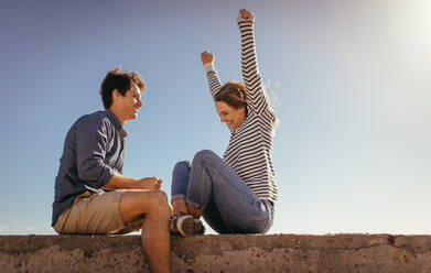 Aufgeregte Frau mit erhobenen Händen, die mit ihrem Freund auf einer Mauer am Meer sitzt. Paar, das an einem sonnigen Tag im Urlaub auf einer Betonmauer sitzt. - JLPSF16727