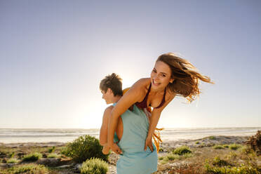 Rückansicht eines Mannes, der seine Freundin auf dem Rücken trägt und zum Meer läuft. Glückliches Paar, das sich an einem sonnigen Tag am Strand vergnügt. - JLPSF16735