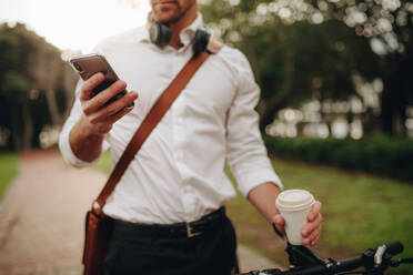 Man checking his mobile phone holding a disposable coffee cup while commuting to office. Businessman using mobile phone while walking to office. - JLPSF16739