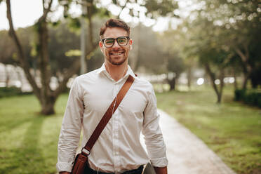 Businessman going to office by walk listening to music on earphones. Happy businessman wearing office bag standing in the street while going to work. - JLPSF16740