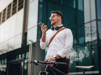 Ein Mann hält sein Fahrrad und spricht über sein Handy, während er ins Büro geht. Ein lächelnder Geschäftsmann spricht über sein Handy, während er am Morgen ins Büro geht. - JLPSF16742