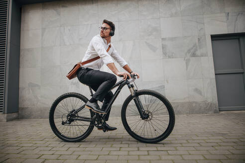 Seitenansicht eines Geschäftsmannes, der mit dem Fahrrad ins Büro fährt, mit Bürotasche und drahtlosen Kopfhörern auf dem Fahrrad. - JLPSF16745