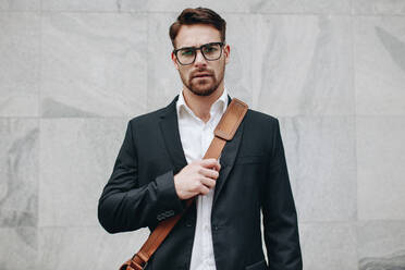 Geschäftsmann mit Bürotasche, der an einer gekachelten Wand steht. Junger Mann mit Brille und Bürotasche im Freien. - JLPSF16747