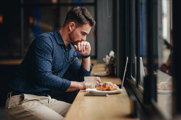 Seitenansicht eines Geschäftsmannes, der in einem Restaurant an einem Laptop arbeitet. Mann sitzt in einem Restaurant mit Essen und Kaffee und arbeitet an einem Laptop. - JLPSF16752