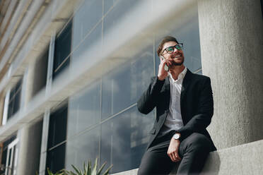 Smiling man talking on mobile phone sitting outside a building. Man taking a break from work talking on cell phone sitting outside his office. - JLPSF16755