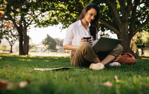 Schöne Frau sitzt auf Rasen im Park, hält einen Kaffee und schaut auf einen Laptop. Junge Geschäftsfrau in legerer Kleidung sitzt im Park mit Kaffee und benutzt einen Laptop. Frau schaut sich ein Video auf dem Laptop an. - JLPSF16757