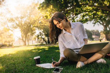 Geschäftsfrau sitzt mit Laptop im Gras im Park und macht Notizen in ihrem Tagebuch. Freiberuflerin arbeitet im Park sitzend. - JLPSF16758