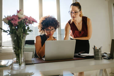 Zwei junge Frauen besprechen das Projekt im Büro über einem Laptop. Geschäftskollegen diskutieren, während sie auf einen Laptop auf einem Schreibtisch vor ihnen schauen. - JLPSF16765