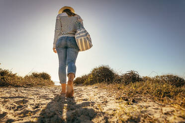 Frau im Urlaub, die mit nackten Füßen auf Sand zum Meer läuft. Frau mit Hut und Tasche, die an einem sonnigen Tag allein am Strand spazieren geht. - JLPSF16781