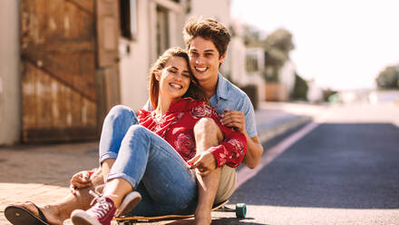 Ein glückliches Paar sitzt zusammen auf einem Skateboard und rollt eine leere Straße hinunter. Ein Mann sitzt auf einem Rollerskateboard mit einer Frau und genießt eine Fahrt im Freien. - JLPSF16787