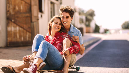 Happy couple sitting together on a skateboard rolling down an empty street. Man sitting on a roller skateboard with a woman enjoying a ride outdoors. - JLPSF16787