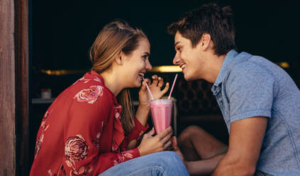 Glückliches Paar, das einen Milchshake mit zwei Strohhalmen trinkt und sich dabei ansieht. Lächelnder Mann und lächelnde Frau bei einem Date, die in einem Restaurant sitzen und einen Milchshake trinken. - JLPSF16789