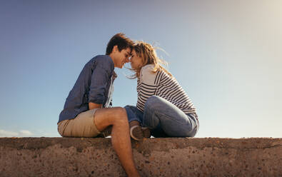 Romantic couple sitting opposite to each other on a sea wall touching their foreheads. Man and woman in romantic mood sitting together at a promenade. - JLPSF16796