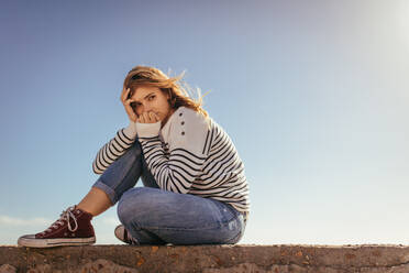 Woman sitting on concrete wall near the sea shore looking away with clear sky in the background. - JLPSF16798