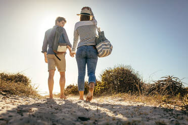 Rückansicht eines Paares, das auf dem Sand spazieren geht und sich an der Hand hält, mit der Sonne im Hintergrund. Mann und Frau, die ihren Urlaub am Strand genießen und mit ihren Taschen spazieren gehen. - JLPSF16801