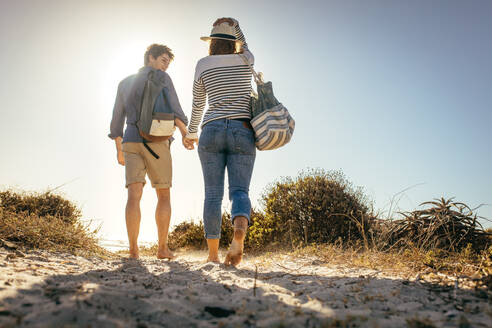 Rear view of a couple walking on sand holding hand with sun in the background. Man and woman enjoying holiday at a beach walking with their bags. - JLPSF16801