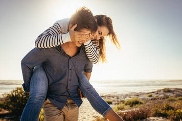 Glücklicher Mann trägt seine Freundin auf dem Rücken am Strand mit der Sonne im Hintergrund. - JLPSF16805