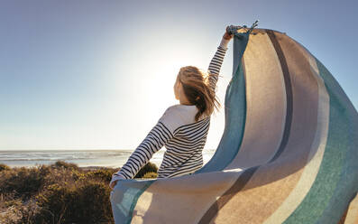 Rückansicht einer Frau, die am Meer steht und ein fliegendes Tuch in beiden Händen hält. Frau hält ein Tuch gegen die Meeresbrise an einem sonnigen Tag. - JLPSF16807