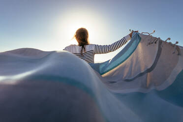 Rückansicht einer stehenden Frau, die ein fliegendes Tuch in beiden Händen hält. Frau hält ein Tuch mit Sonnenfackel im Hintergrund. - JLPSF16809