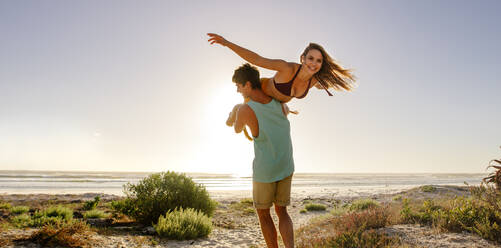 Glückliches Paar, das sich an einem sonnigen Tag am Strand vergnügt. Die Frau breitet ihre Arme aus, während sie von ihrem Freund getragen wird. - JLPSF16811